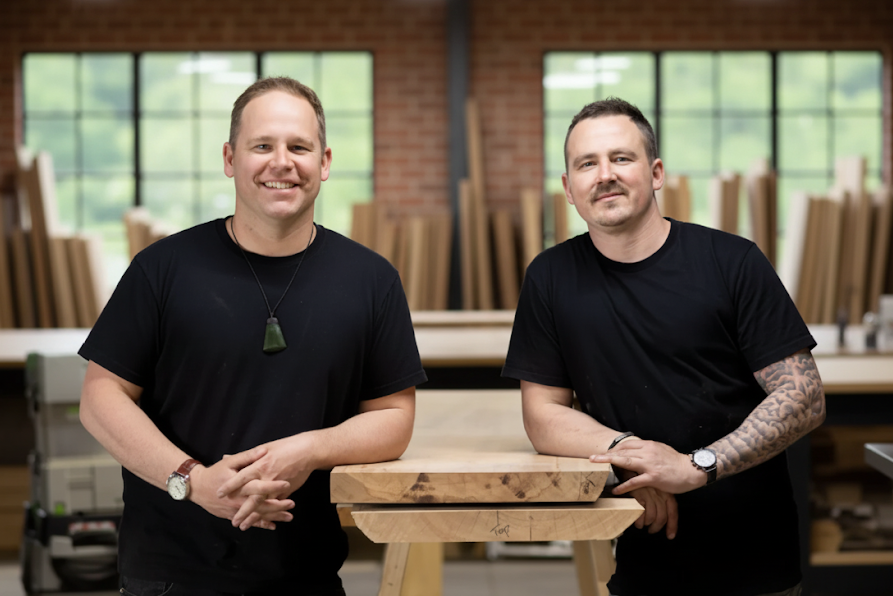 Two men standing in a workshop with wooden boards and tools in the background