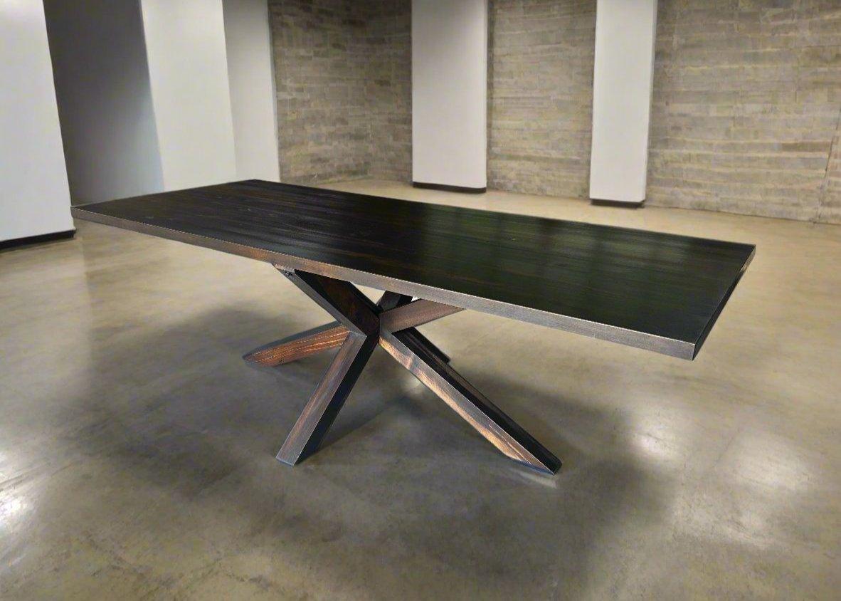 A round solid wood dining table with a sculptural X-shaped wooden pedestal base, displaying rich natural wood tones, positioned as a centrepiece in a modern dining room.