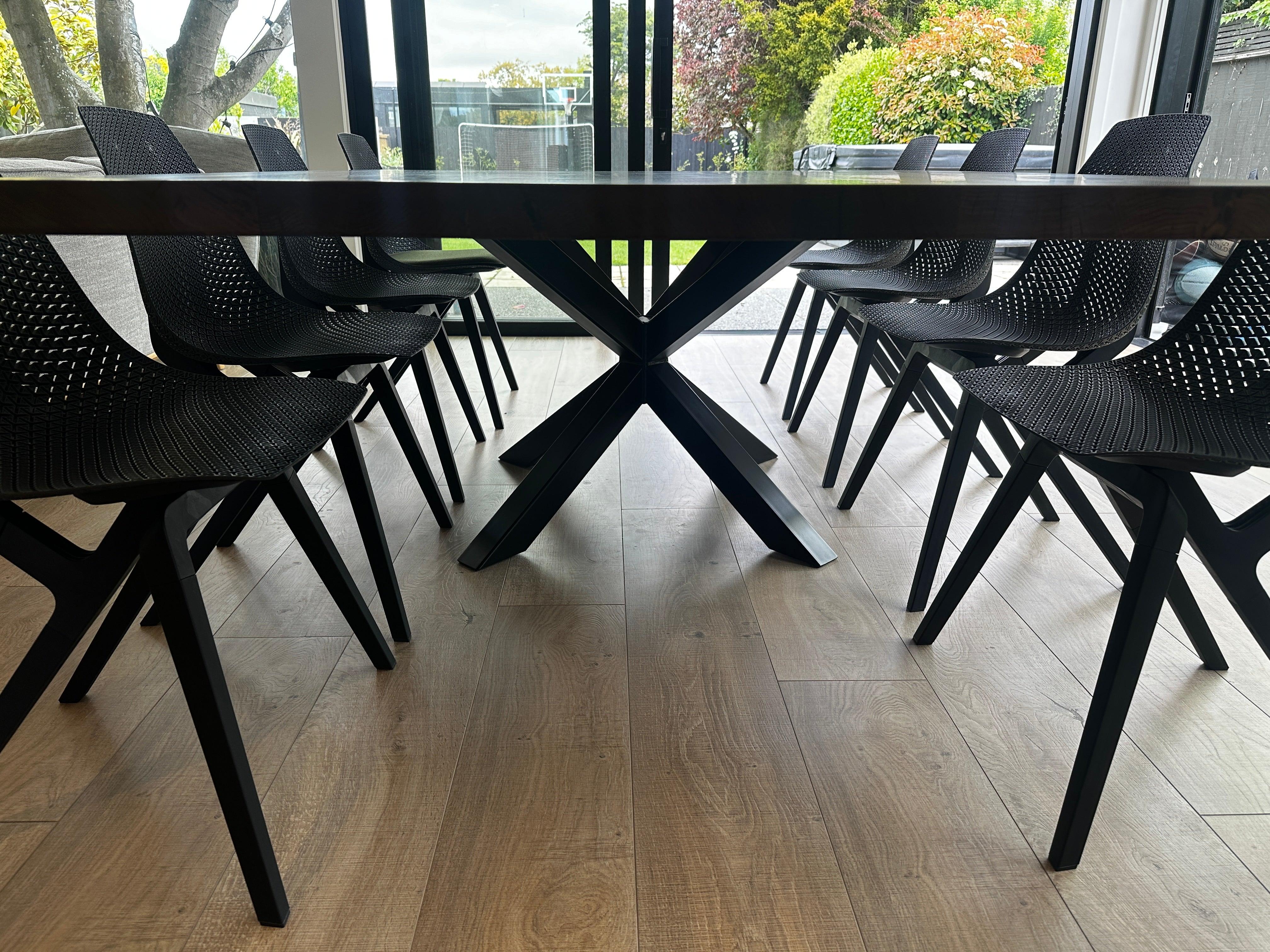 A modern dining table with a thick solid wood top and a black steel X-shaped pedestal base, surrounded by dining chairs in a contemporary home setting