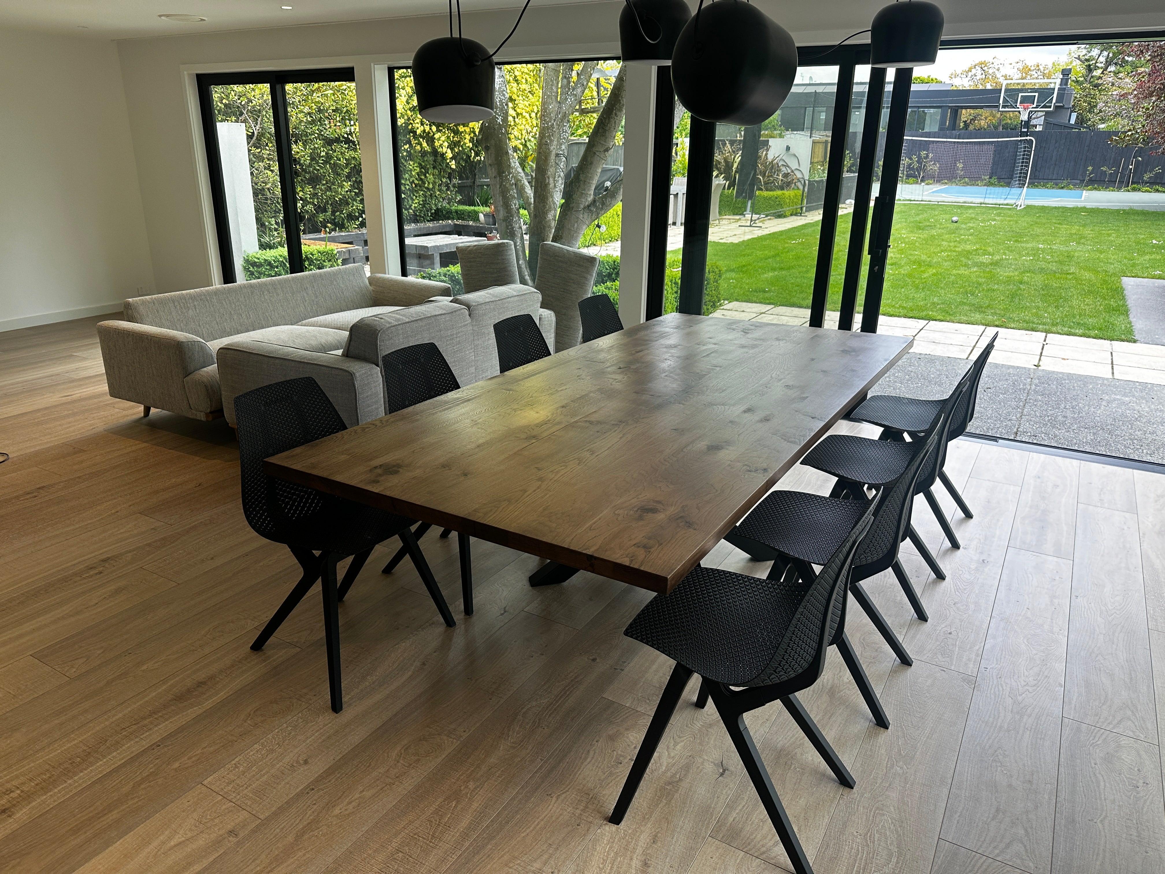 A modern dining table with a thick solid wood top and a black steel X-shaped pedestal base, surrounded by dining chairs in a contemporary home setting