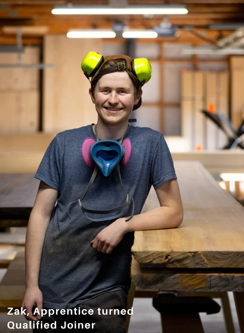 Workshop worker wearing earmuffs and a respirator around the neck, leaning on a wooden surface