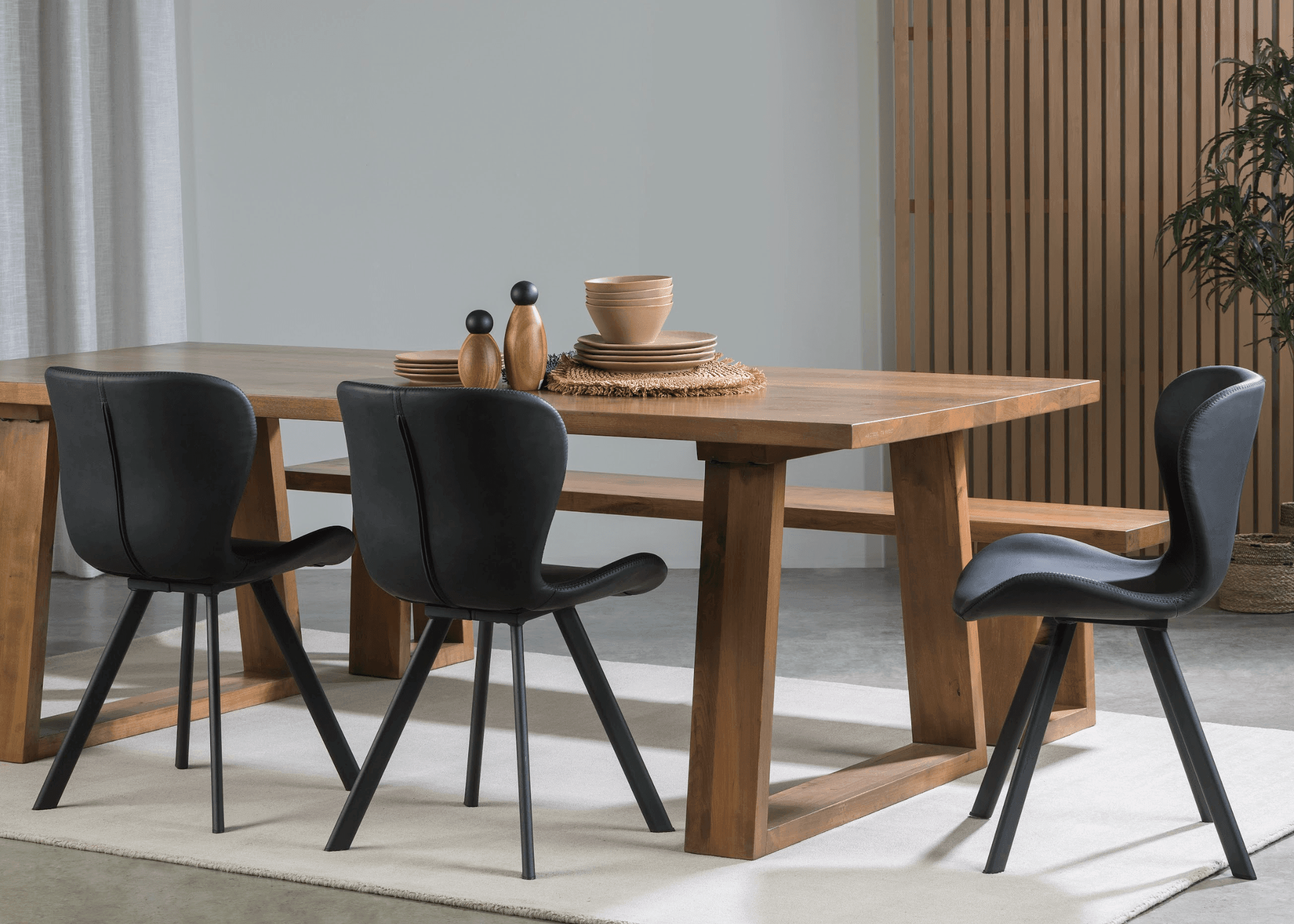 A solid wood dining table featuring uniquely angled wooden legs and a matching bench, showcasing natural timber grain in an airy modern dining space.
